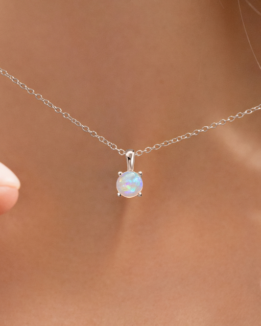 A close-up of a delicate silver chain necklace with a round iridescent opal pendant, worn on a model's neck.